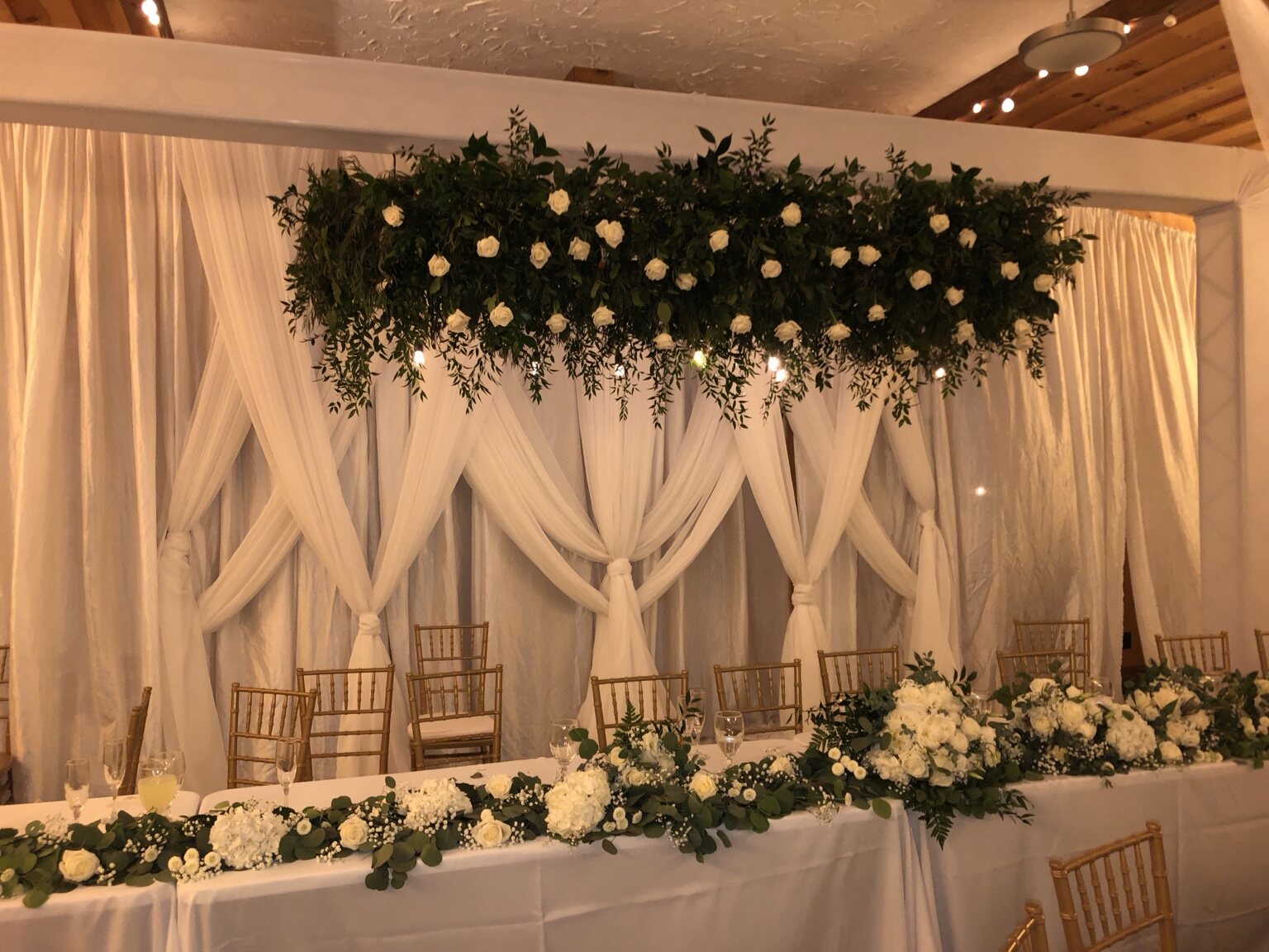 Wedding head table framed by white drapery, greenery and ivory flowers